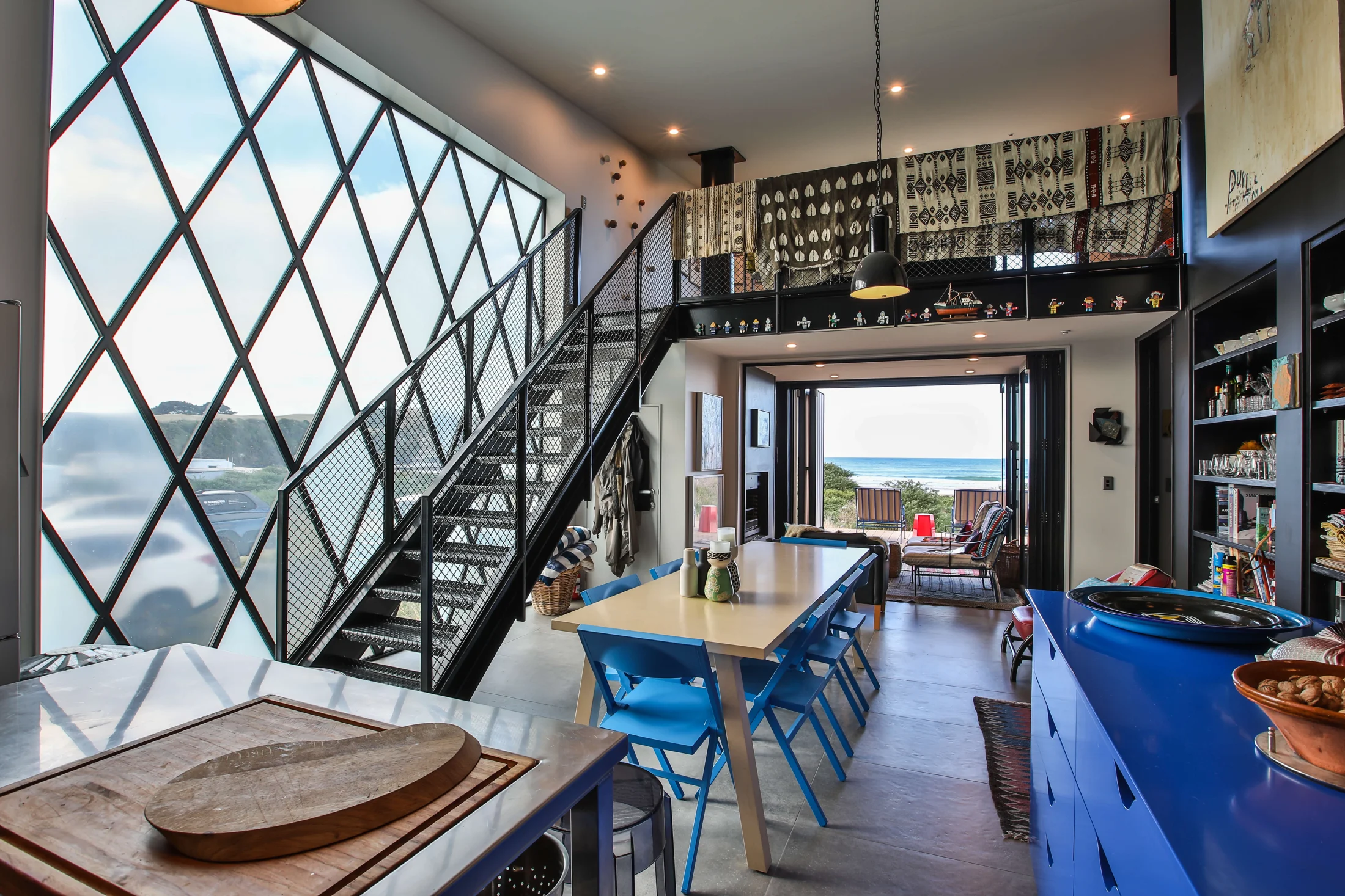 A coastal interior with a sculptural steel staircase, blue chairs and ocean views